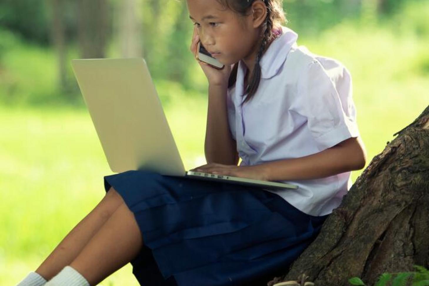 Girl sitting at the base of a tree on a laptop talking on the phone.