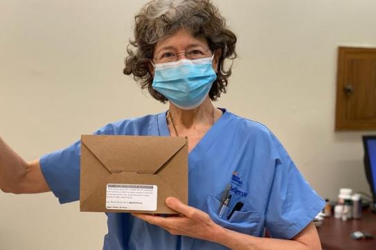 Healthcare worker in scrubs and a mask holds a boxed meal, giving a thumbs-up